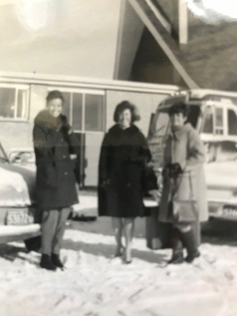 A blurry photo of three young women wearing winter clothing, standing in front of a bus, some cars and a small building, smiling.